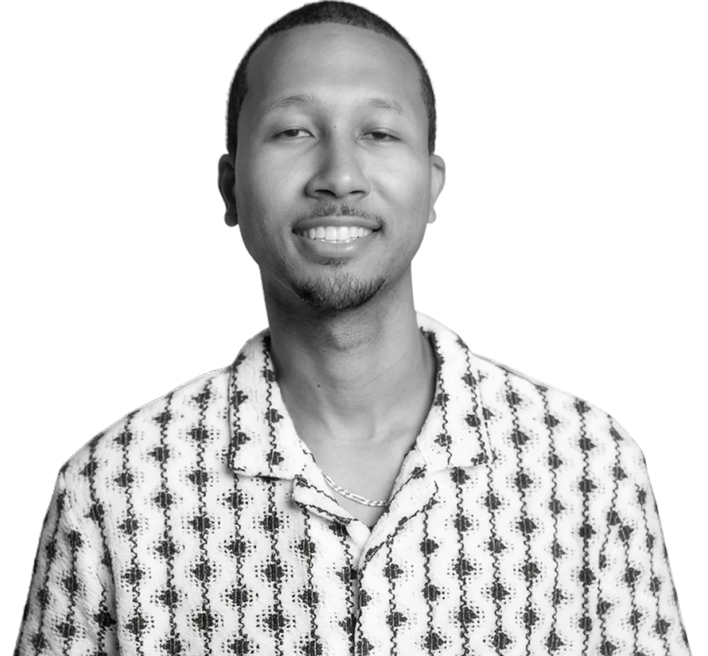 Black-and-white portrait of a smiling man wearing a patterned shirt and chain necklace against a bright yellow background.